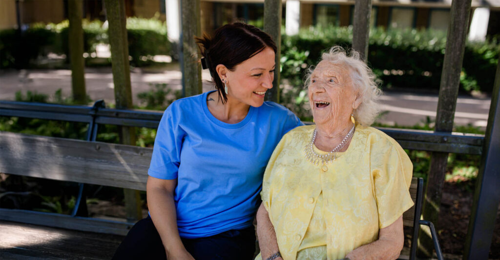 An employee at Ambea is sitting with a laughing elderly woman on a park bench.